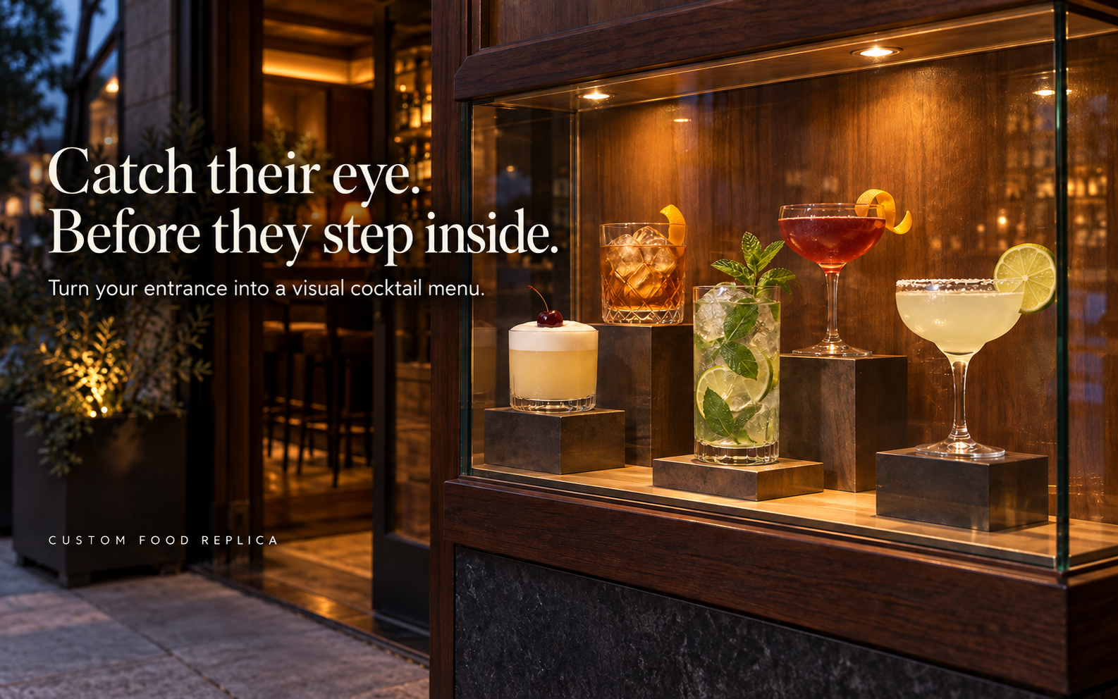 Street facing bar entrance with a warm lit case of five cocktail replicas on risers